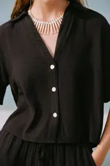 Camisa negra de cuello solapa con escote pico y manga corta. Detalle de nido de abeja en espalda. Cierre frontal con botones.