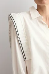 Camisa de color blanco con cuello clásico, cierre frontal de botones y mangas largas con puños. Presenta detalles de bordado geométrico en contraste a lo largo de los hombros y la parte superior de la espalda.