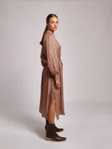 Vestido midi de satén con estampado geométrico en tonos marrones y naranjas. Presenta cuello mao, mangas largas abullonadas con puños ajustados y un cinturón a tono con hebilla metálica que marca la cintura.