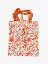 Tote bag plegable con estampado floral en tonos rojos, naranjas y azules sobre un fondo beige. Tiene asas de tela color naranja.