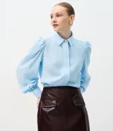 Camisa de crepé color blanco, con cuello inglés, manga larga abullonada con puños abotonados y cierre frontal oculto con botón diferenciado en el cuello.