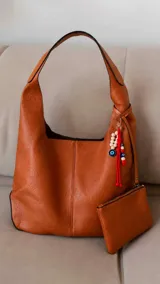 Cartera tipo hobo de cuero sintético color suela, con interior espacioso y costuras decorativas frontales. Incluye asa de hombro fija, monedero rectangular colgante a juego y un llavero amuleto con cuentas de madera, ojos turcos y detalles en rojo.