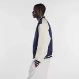Campera estilo varsity con cuerpo azul marino y mangas en color beige. Presenta cierre frontal con botones a presión, cuello, puños y ruedo acanalados con rayas decorativas, y logo de la marca bordado en el pecho.
