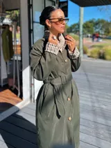Trench reversible de corte clásico con cinturón ajustable. Un lado presenta un diseño liso en color verde militar, mientras que el reverso cuenta con un estampado a cuadros en tonos neutros. Posee cierre frontal con botones y cuello camisero.