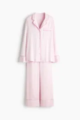Pijama de dos piezas en tejido fluido de satén color rosado con ribetes en color contrastante. Camisa de manga larga con cuello abierto, botones adelante y bolsillo superior abierto, junto con un pantalón de pierna amplia.
