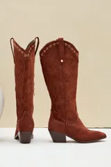 Bota texana de gamuza color marrón, con taco y puntera fina. Presenta bordados decorativos en el cuerpo de la caña y detalles de tachas metálicas en la parte superior y en la costura inferior.