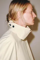 Trench corto color marfil de algodón, con cuello camisero, solapa cortaventos, mangas largas amplias con puños abotonados y cierre cruzado con un botón visible.