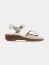 Sandalia de cuero con tiras ajustables de velcro en el empeine y el talón. Presenta un diseño bicolor en tonos dorado y beige, con una suela de goma texturizada de aspecto natural.
