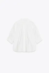Camisa blanca de algodón con cuello solapa, detalle de pliegues en el delantero y mangas tres cuartos abullonadas terminadas en puño. Cierre frontal con botones ocultos.