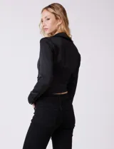 Blusa negra de satén, marca Allie Rose, con cuello con solapa y escote en V. Diseño cruzado con lazo de ajuste lateral y manga larga con pretina, ojal y botón.