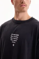 Remera oversize de manga corta en color negro con efecto acid wash. Presenta un pequeño estampado frontal con texto en inglés y un isotipo de flor en la parte superior del pecho.