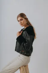 Chaqueta de cuero Napa negra con cuello y puños de lana sintética negra. Cierre frontal con cremallera y detalles de botones metálicos en la parte inferior.