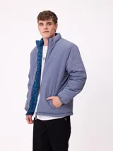 Campera reversible de nylon con cuello alto y cierre frontal. Un lado es de color gris liso con logo bordado, mientras que el otro lado presenta un estampado abstracto en tonos azules. Cuenta con bolsillos laterales y puños elásticos.