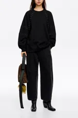 Sudadera negra de corte oversize, confeccionada en algodón, con escote redondo y mangas largas tipo ranglán. Presenta detalles de remaches metálicos distribuidos a lo largo de las costuras de las mangas.