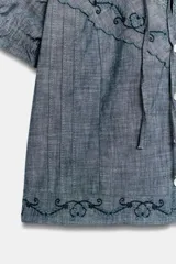 Camisa de cuello redondo con lazada frontal, manga corta abullonada con lazos ajustables y cierre frontal con botones. Presenta detalles de bordados y encaje a tono sobre una base de tela tipo chambray.