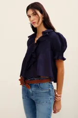 Camisa azul marino de algodón, con mangas cortas abullonadas y puños fruncidos con elástico. Presenta un amplio escote en V adornado con volados con detalles bordados y ojales a lo largo de la botonera frontal.