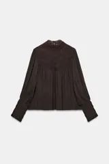 Blusa de cuello subido y manga larga, color marrón oscuro, con detalles de bordados combinados a tono en el canesú y los puños anchos. Cierre en la espalda con abertura y botones.