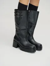 Bota alta negra de cuero vacuno wax, con punta cuadrada, suela de goma con plataforma y taco de madera. Cuenta con detalle de dos hebillas metálicas en la parte superior de la caña.