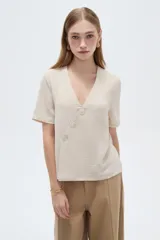 Blusa de poliéster color beige, con cuello en V y diseño cruzado con tres botones en diagonal. Cuenta con mangas cortas y calce holgado.