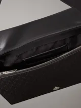 Cartera de mano Calvin Klein con correa ajustable para el hombro. El material exterior es de piel sintética negra con un patrón texturizado de monograma repetido. Presenta una solapa con cierre magnético y un logo metálico plateado en la parte frontal.
