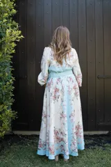 Vestido largo estampado con motivos florales en tonos pastel y detalles de cadenas doradas. Presenta cuello mao, cierre frontal con botones variados, mangas largas con puños elásticos y un cinturón a tono con hebilla dorada que realza la cintura. El ruedo del vestido tiene una franja de color celeste.