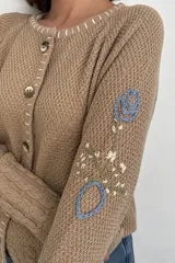 Saco de punto color beige, confeccionado en algodón, con cuello redondo, mangas largas y cierre frontal con botones. Presenta detalles bordados con diseño floral en las mangas y ribetes con rayas azules y blancas en la botonera y el cuello.