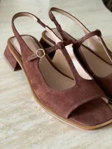 Sandalia de gamuza color marrón, con diseño tipo T-strap, taco cuadrado bajo y tira ajustable al talón con hebilla metálica dorada.