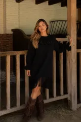 Vestido de punto negro con cuello y puños decorados con volados. Presenta mangas largas y un ruedo terminado en flecos largos.