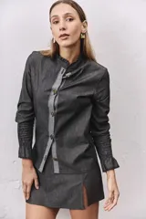Camisa de jean gris oscuro con cuello mao, cierre frontal con botones metálicos y una solapa en contraste de color gris claro. Presenta mangas largas con puños elásticos fruncidos y volados en los extremos.