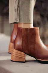 Botín de cuero negro, caña corta, con punta cuadrada y cierre lateral. Presenta un tacón de madera de altura media y detalles en charol con textura de cocodrilo en el talón.