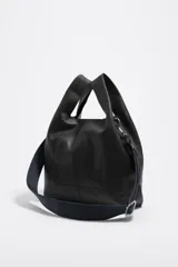 Bolso de mano mediano de piel vacuna, color negro, con estructura blanda y forma de cubo o 'paper bag'. Posee asas de mano cortas y una correa ajustable y extraíble de tela azul marino. El logo de Bimba y Lola está grabado en la parte inferior frontal.