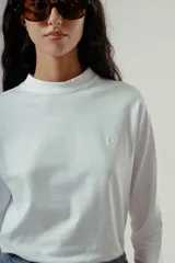 Camiseta de manga larga, cuello redondo, con rayas horizontales marrones sobre fondo blanco. Presenta un pequeño logo bordado en rojo en el pecho.