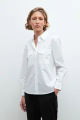 Camisa blanca de manga larga con cuello clásico y cierre frontal con botones. Presenta dos bolsillos plaqué en el pecho.