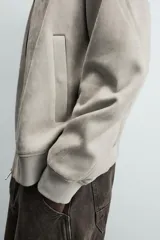 Cazadora bomber negra de corte regular fit, confeccionada en tejido efecto ante. Cuenta con cuello, puños y bajo en rib elástico, manga larga, bolsillos de vivo en la cadera y un bolsillo interior. Cierre frontal con cremallera.