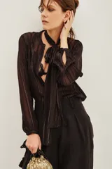 Camisa de manga larga en color negro con rayas verticales metalizadas. Presenta un diseño con lazo integrado en el cuello y puños abotonados. Confeccionada en tejido semitransparente con caída fluida.