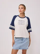 Remera bicolor Rusty de corte holgado y estilo deportivo, con cuerpo blanco y mangas raglán azules con doble raya blanca. Presenta estampado frontal con el texto 'RUSTY 85'.