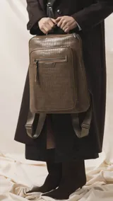 Mochila marrón con textura símil cocodrilo, compartimento principal con cierre, amplio bolsillo frontal con cierre y correas ajustables de tela.