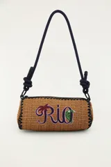 Bolso tipo bandolera de formato cilíndrico, confeccionado con material trenzado en color tostado, con detalle de bordado frontal que dice 'Rio' acompañado de una palmera y un tucán. Cuenta con una correa larga de cuerda oscura con nudos ajustables y cierre superior.