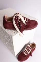 Zapatillas urbanas color tostado con textura geométrica en el cuerpo y detalles de gamuza en la puntera y laterales. Presentan un diseño distintivo con cordones anchos tejidos en tonos tostado y blanco, y un accesorio metálico con borla de gamuza en el empeine.