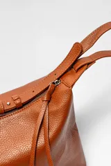 Bolso de hombro tipo hobo, confeccionado en cuero texturizado color marrón. Presenta un diseño minimalista con cierre superior, correa ajustable y detalle de tiras decorativas en el lateral.