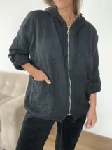 Campera fina de algodón color negro con capucha, cierre frontal y detalles tipo broderie. Presenta un corte holgado, mangas con opción de remangar y tiras regulables en la parte inferior.