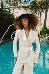 Blazer de corte estructurado en color beige, con cuello solapa y manga larga. Presenta un diseño cruzado con cierre mediante lazada del mismo tejido y un distintivo aplique metálico dorado en el frente.