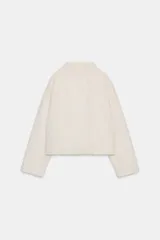 Cazadora de textura suave efecto pelo, color crudo, con cuello subido y cierre frontal cruzado con botones. Presenta mangas largas y bolsillos laterales ocultos en las costuras.
