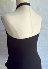 Vestido corto negro, sin mangas, con escote halter que se ajusta con una argolla oscura en el centro del pecho. La parte superior tiene un drapeado suelto y la falda es ajustada.