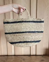 Bolso tipo cartera de mano confeccionado en fibras naturales trenzadas, con diseño estructurado y base redondeada. Presenta franjas horizontales en color negro y doble asa superior corta trenzada.