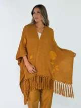 Ruana de algodón color crudo, tejida en punto abierto con detalles de flores bordadas en relieve y flecos en el borde inferior.