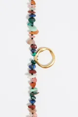 Collar estilo bolo o lariat, compuesto por piedras semipreciosas de diferentes colores y formas irregulares, ensartadas en dos hebras que se unen mediante un anillo central dorado. Las hebras cuelgan libremente y terminan en tubos metálicos dorados.