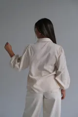 Camisa de color beige con mangas abullonadas y puños ajustados. Presenta cuello clásico, cierre frontal con botones y detalles bordados sutiles en las mangas.