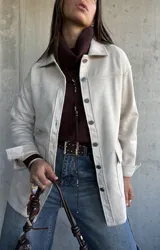 Chaqueta tipo sobrecamisa color beige, con cierre frontal de botones metálicos, cuello clásico y bolsillos frontales. Presenta un diseño de corte recto y mangas largas con puños abotonados.
