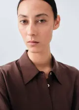 Camisa de seda de corte oversize, color marrón chocolate, con cuello camisero, mangas largas con puños abotonados y cierre frontal de botones ocultos.